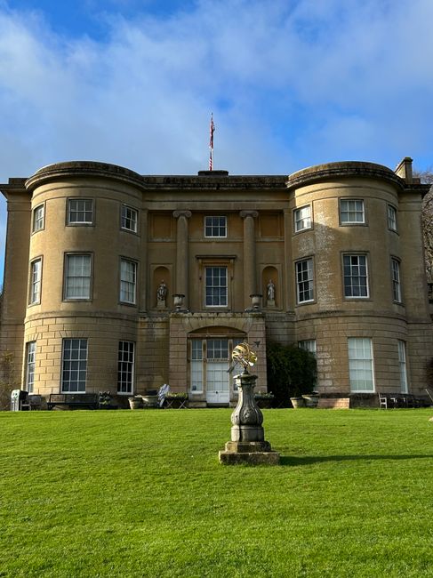 A grand, neoclassical-style building with a symmetrical facade, featuring large windows and decorative columns at the entrance. In the foreground, there is a well-maintained, lush green lawn with a de